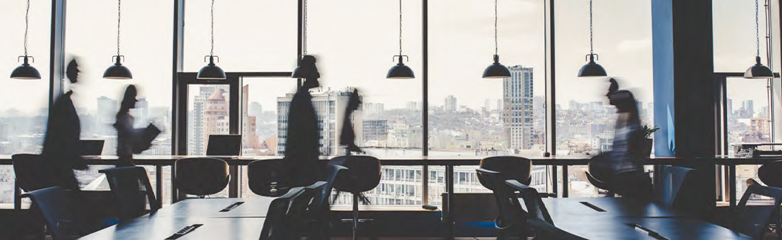 Academic research office with large windows facing city skyline with blurred figures walking between desks and pendant lights
